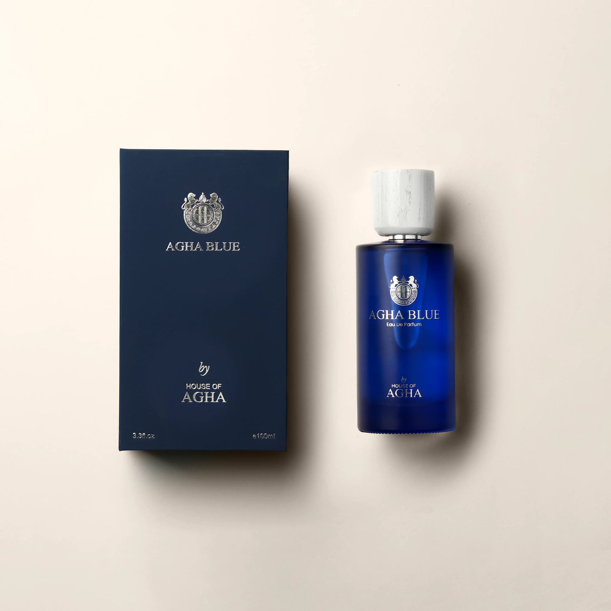 A combined image featuring a deep blue bottle of Agha Blue Eau de Parfum by House of Agha with a white cap and elegant silver branding, placed next to its matching deep blue box displaying the brand logo and name in silver, with a capacity of 3.3 fl oz (100ml), set against a plain background.