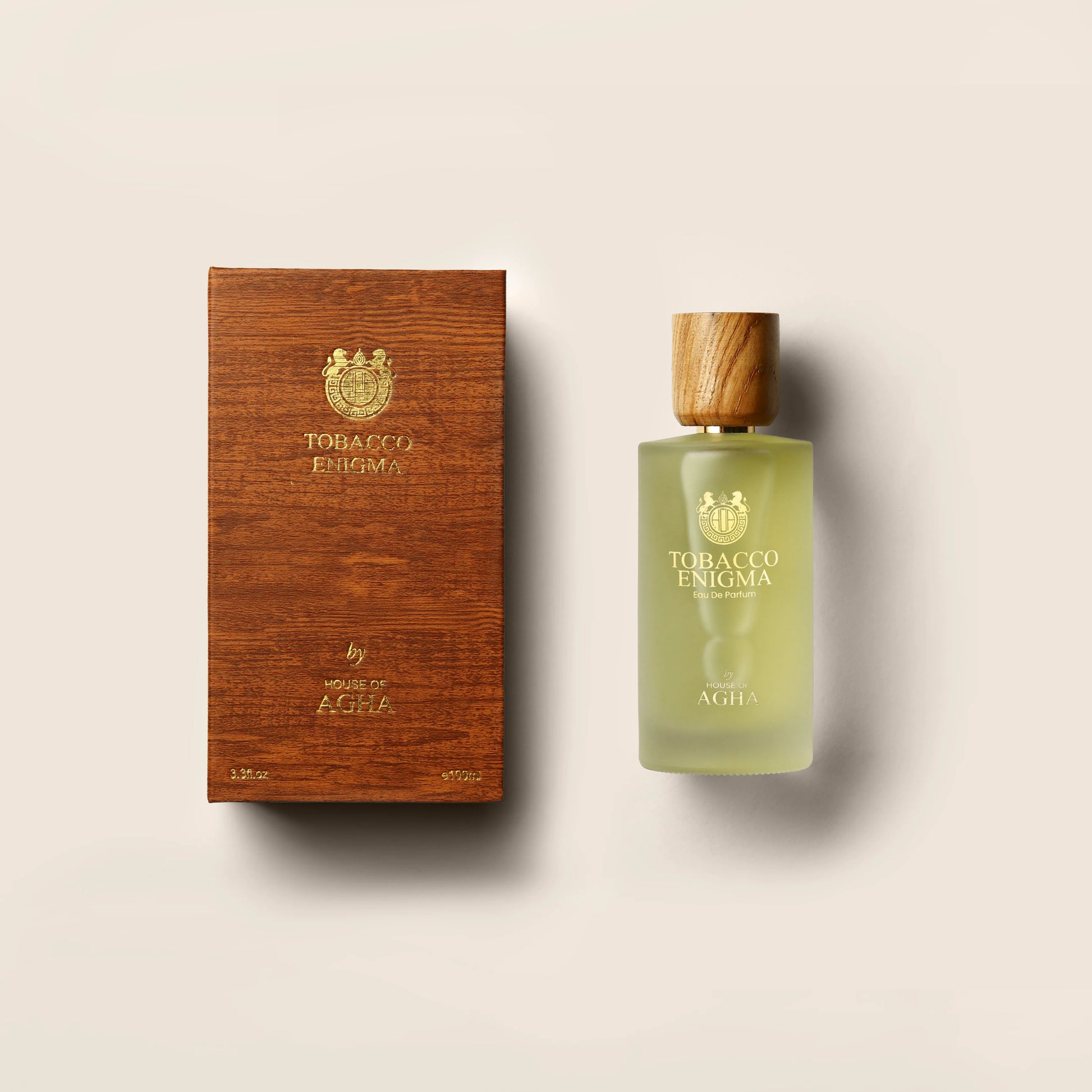 A combined image featuring a light green bottle of Tobacco Enigma Eau de Parfum by House of Agha with a wooden cap, placed next to its matching wooden-textured box with gold branding, set against a plain background.
