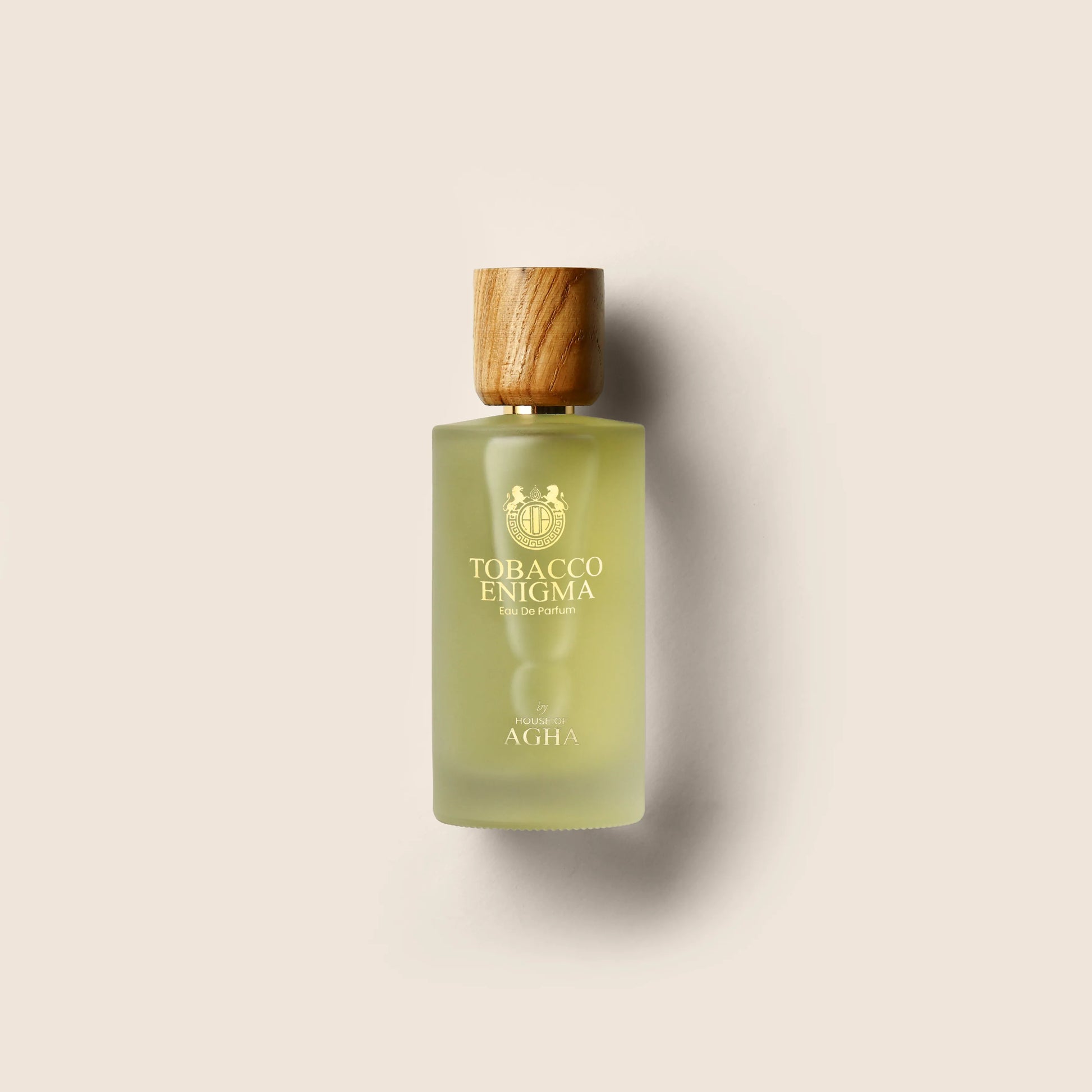 A bottle of Tobacco Enigma Eau de Parfum by House of Agha, featuring a light green glass bottle with a wooden cap and elegant gold branding, placed against a plain background.