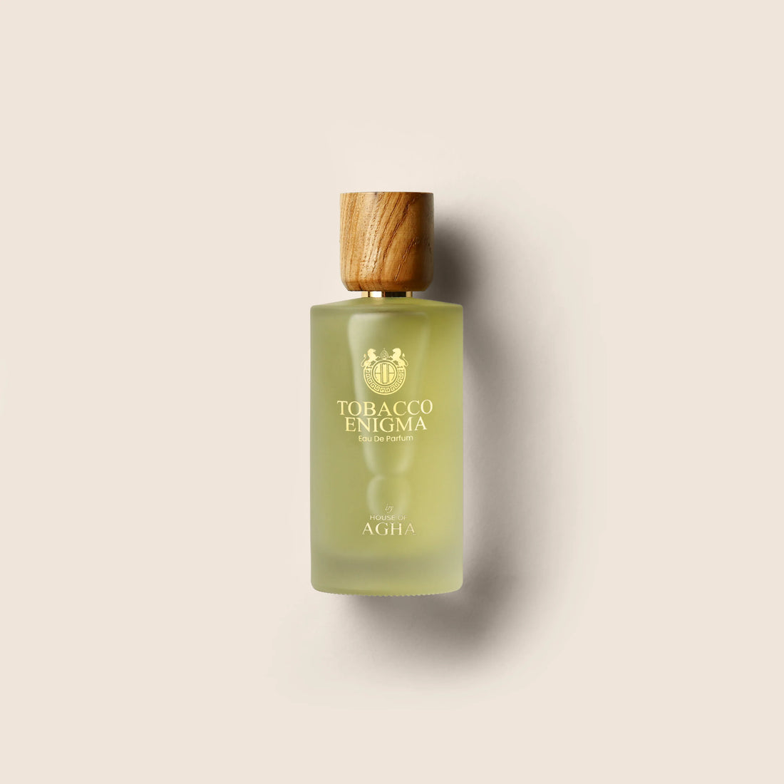 A bottle of Tobacco Enigma Eau de Parfum by House of Agha, featuring a light green glass bottle with a wooden cap and elegant gold branding, placed against a plain background.